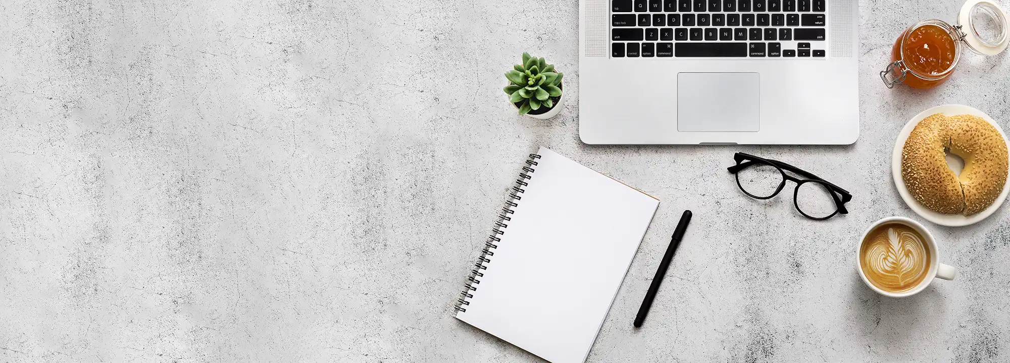 Laptop, notebook, cappuccino, bun, and jam on a desk, representing the clear terms and values of Delica Recipes.