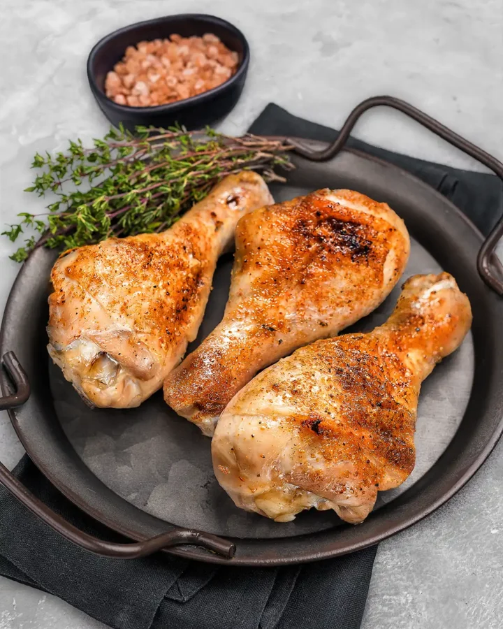 Close-up of crispy baked chicken drumsticks with fresh thyme and pink salt, showing crisp golden skin and and juicy roasted meat.